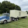 Side view of a blue semi-truck hauling a beige containerized Caterpillar generator enclosure on a triple-axle trailer.