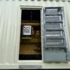 This image shows the breaker access door of a Caterpillar 3516C containerized generator enclosure in the open position. The exterior of the enclosure features ventilation louvers for cooling airflow, while the hinged door includes an integrated fold-out ladder step system for safe operator entry. Through the access opening, the generator circuit breaker and control distribution panels are clearly visible inside, providing convenient access for monitoring, maintenance, and electrical connection operations. This design ensures secure, weather-protected housing while maintaining operator safety and serviceability.