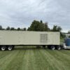 Full left-side view of containerized Caterpillar generator enclosure mounted on a tri-axle trailer, connected to a blue semi-truck.