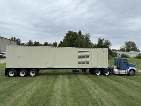 Full left-side view of containerized Caterpillar generator enclosure mounted on a tri-axle trailer, connected to a blue semi-truck.