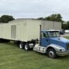 Side view of containerized Caterpillar generator enclosure mounted on a tri-axle trailer, connected to a blue semi-truck.